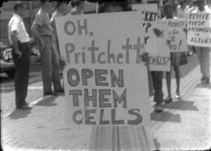 An individual holds a sign with the phrase, "Oh, Pritchett open them cells."